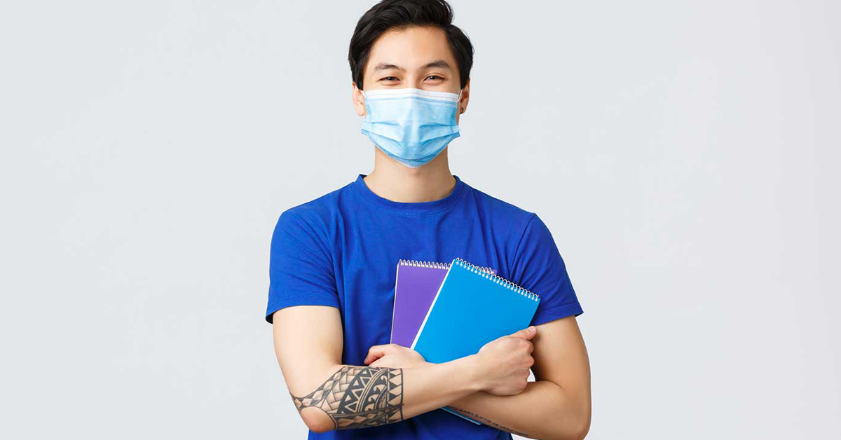 no centro da imagem, jovem amarelo usando máscara e segurando 2 cadernos, um azul, outro roxo. Ele veste camiseta azul e tem tatuagens nos antebraços.