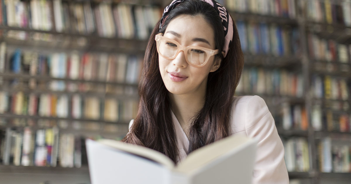 Mulher lendo livro. Ao fundo há uma estante repleta de livros.