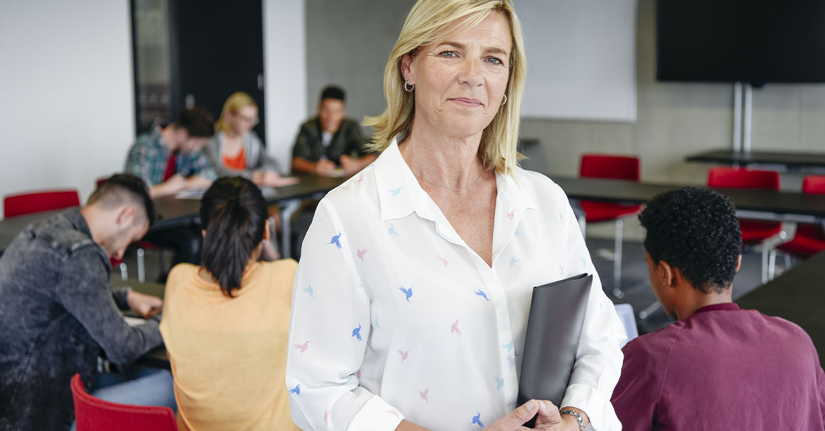 Professora branca., loira em pé em uma sala de aula.