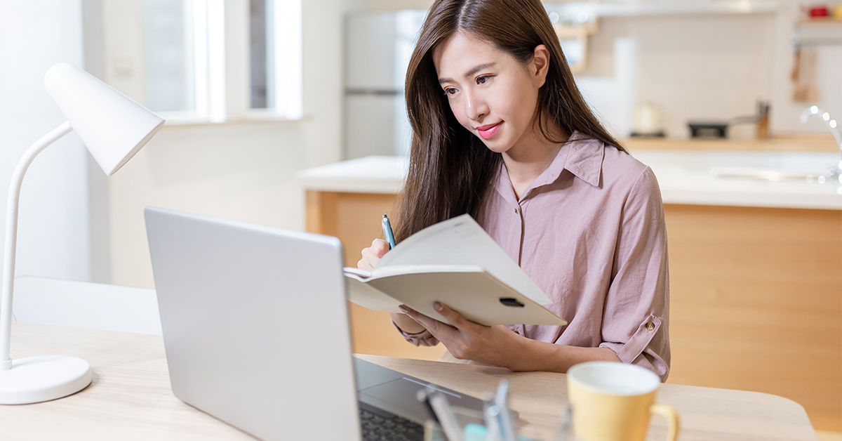 Mulher estudando a distância. Ela, que tem cabelo liso e veste camisa rosa, está sentada diante um computador, com um livro e uma caneta em mãos.