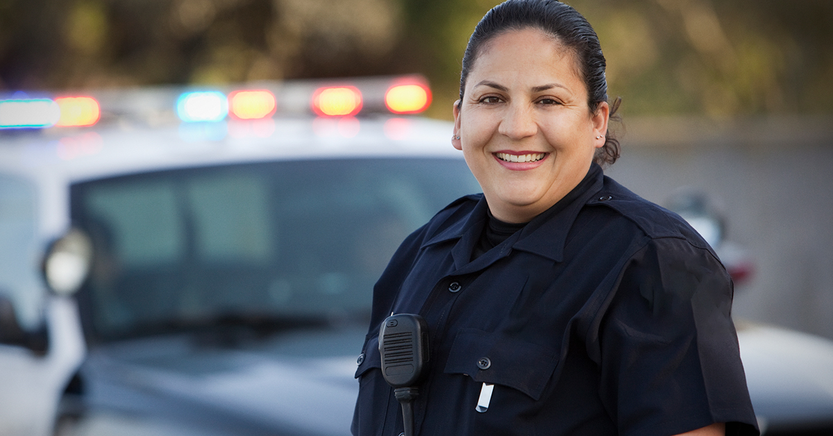 Mulher branca vestida com uniforme de polícia. Ao fundo da imagem, há um carro de polícia, com o giroflex ligado.