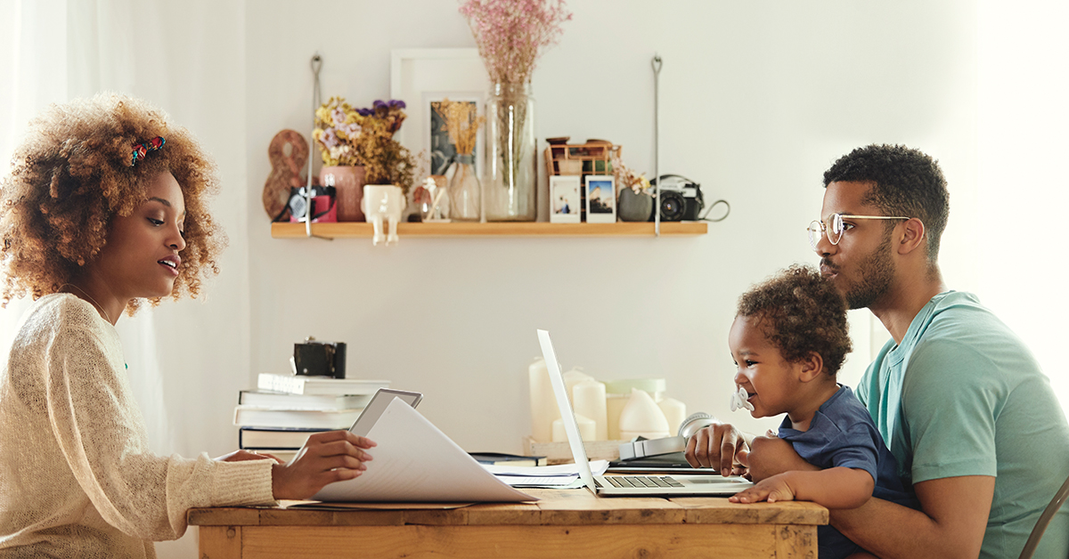 Em home office, à esquerda, mulher negra sentada à mesa com um livro em mão. Ela veste blusa branca. A sua frente, uma criança negra está sentada no colo de um homem negro. Eles estão mexendo em um notebook. A criança veste camiseta azul e está com uma chupeta em boca. Já o homem veste camiseta verde e usa óculos de grau. Na mesa, que está ao centro da imagem, há livros empilhados. Acima, há uma estante repleta de objetos de decoração.