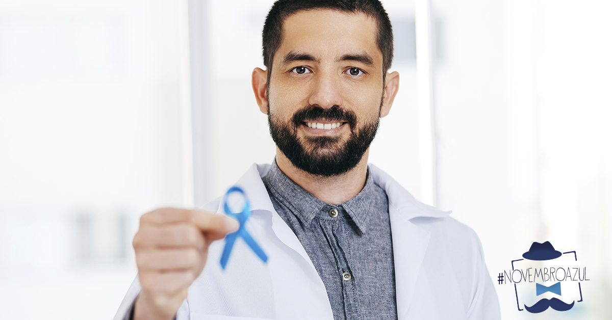 Ao centro da imagem homem branco sorrindo. Ele veste camisa cinza e jaleco branco e está segurando a fita do novembro azul em uma de suas mãos.