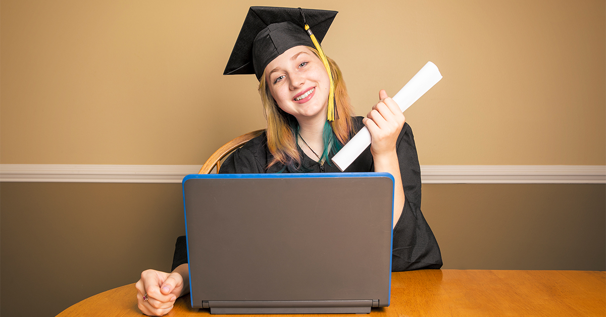 mulher branca e loira vestindo beca, com um diploma em mão. Ela está sentada na frente de um notebook.