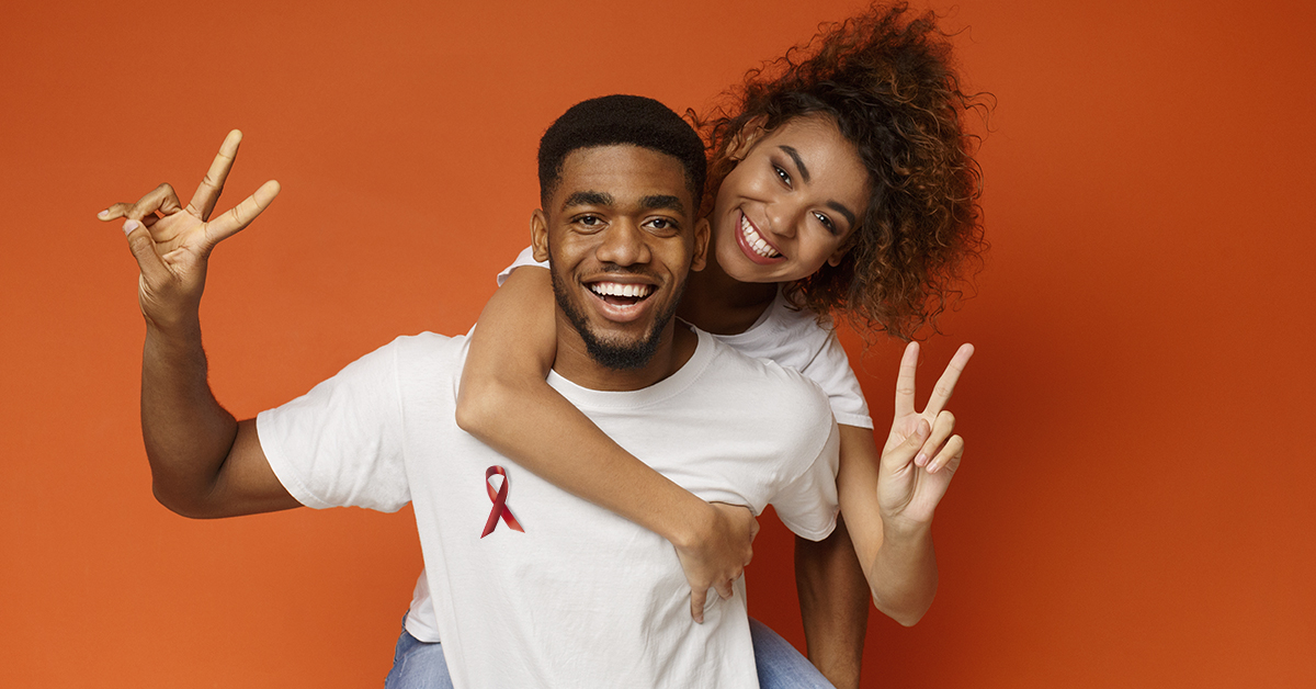 no centro da foto um casal heterossexual negro. O homem veste camiseta branca e está fazendo o número 2 com os dedos de ambas as mãos. A mulher está por trás, abraçada no pescoço dele. Ela está de camiseta branca e calça jeans.