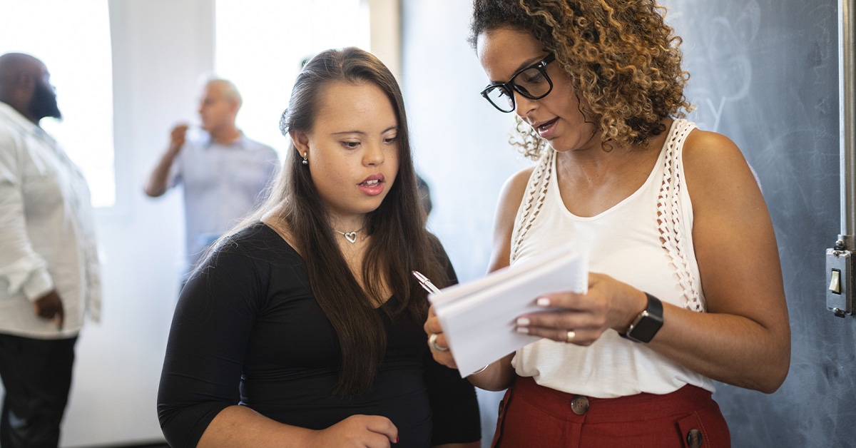 à frente da imagem, mulher negra escreve com uma caneta em um caderno, em mão. Ela, que usa óculos de grau, veste blusa branca e calça vermelha, conversa com jovem com síndrome de down, que veste roupa preta. Atrás da mulher há um quadro verde. Mais ao fundo, há pessoas conversando. A pauta é mercado de trabalho.