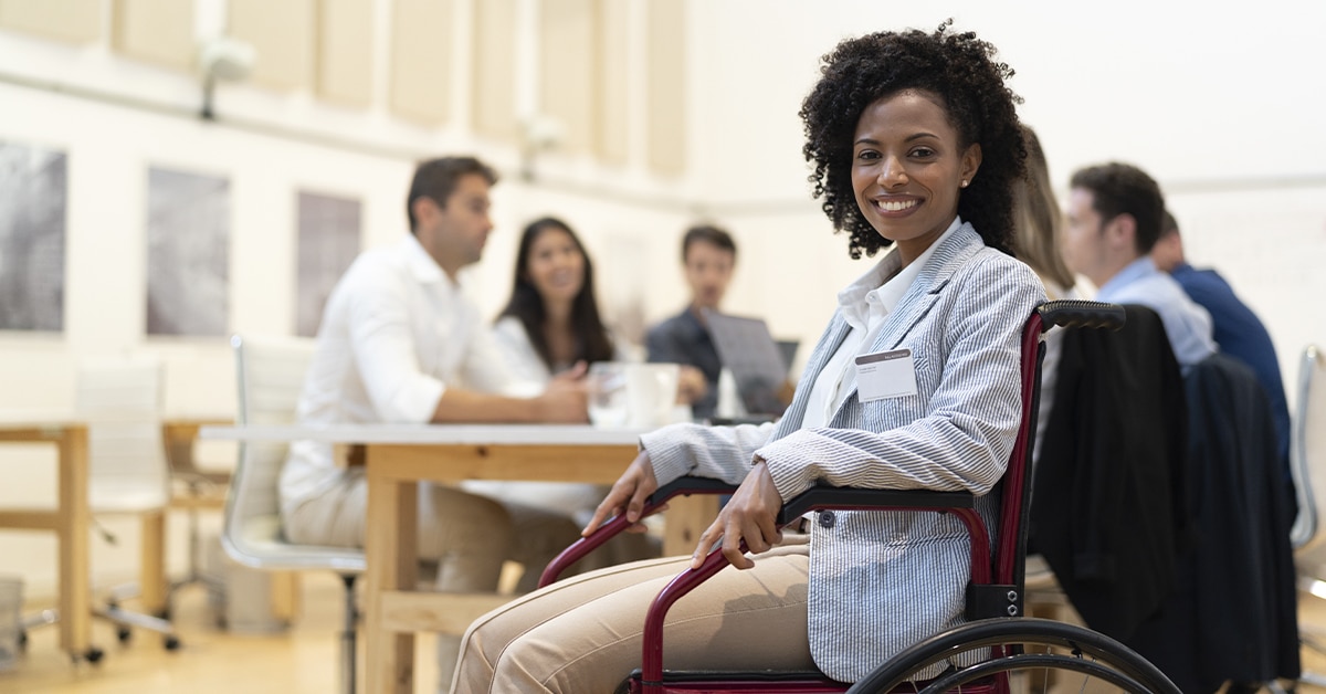 mulher negra e cadeirante à frente e à direita da imagem. Ela, que está sorrindo para a câmera, veste calça social bege, camisa branca e paletó listrado azul e branco. Ao fundo, há quatro pessoas sentadas ao redor de uma mesa. Entretanto, o fundo está bem desfocado.