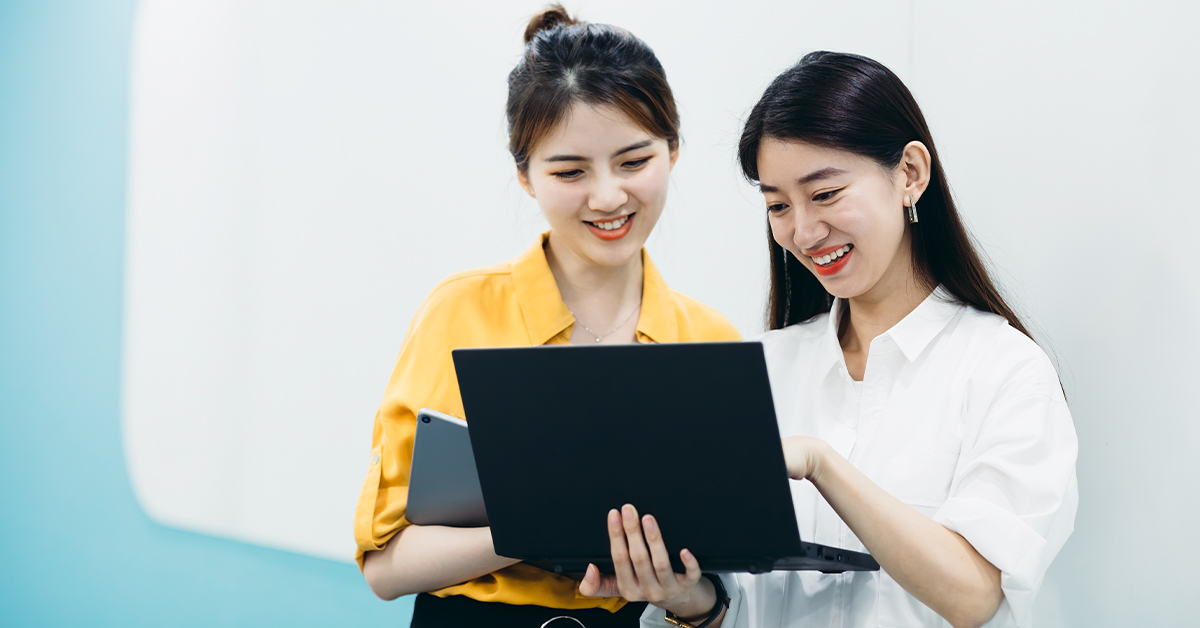 duas mulheres amarelas conversam ao olharem para um notebook. À esquerda, está mulher com camisa amarela e cabelo preso em coque, segurando um tablet no braço, enquanto a sua direita a outra mulher está de camisa branca e cabelo solto. ambas sorriem.