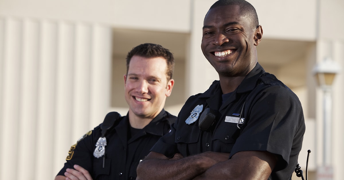 dois policiais sorrindo. À frente, há o policial negro e ao fundo, um policial branco. Os dois estão de lado, levemente inclinados para frente.