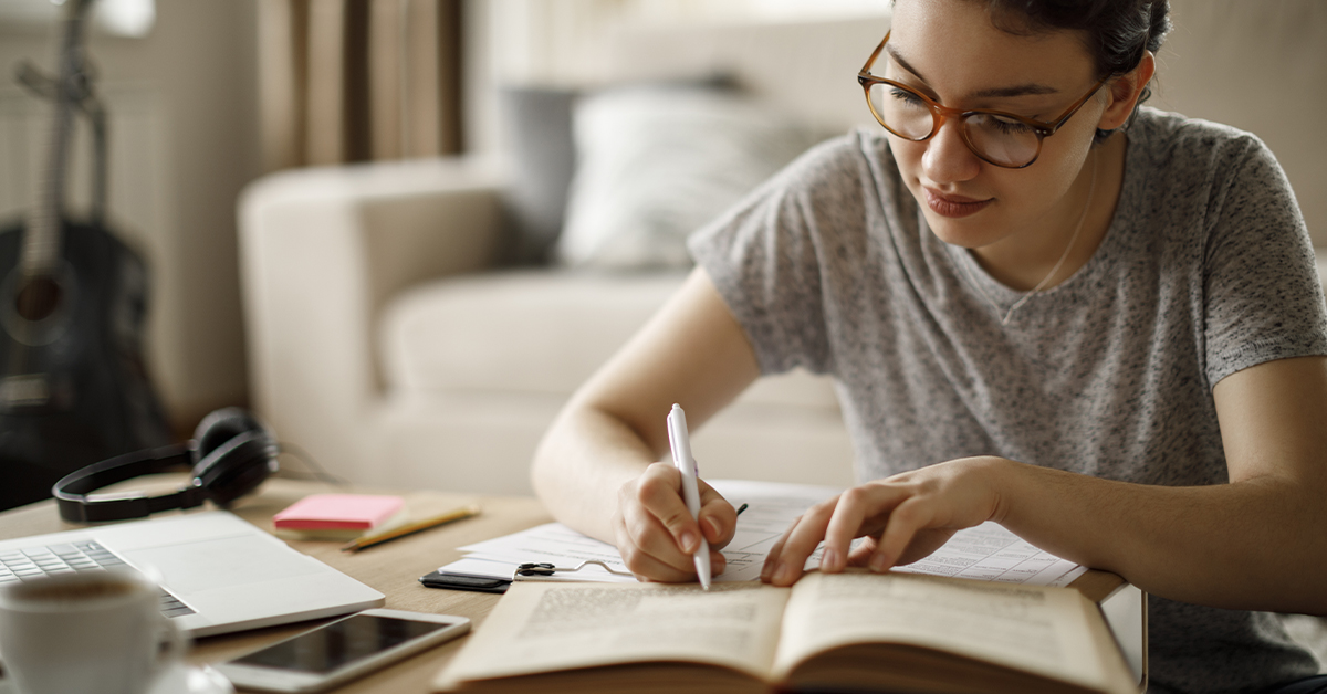 mulher parda estudando. Na mesa há caderno, folhas, notebook, celular e headphones. Ela está sentada no chão e escrevendo em uma folha sobre uma mesa de centro.