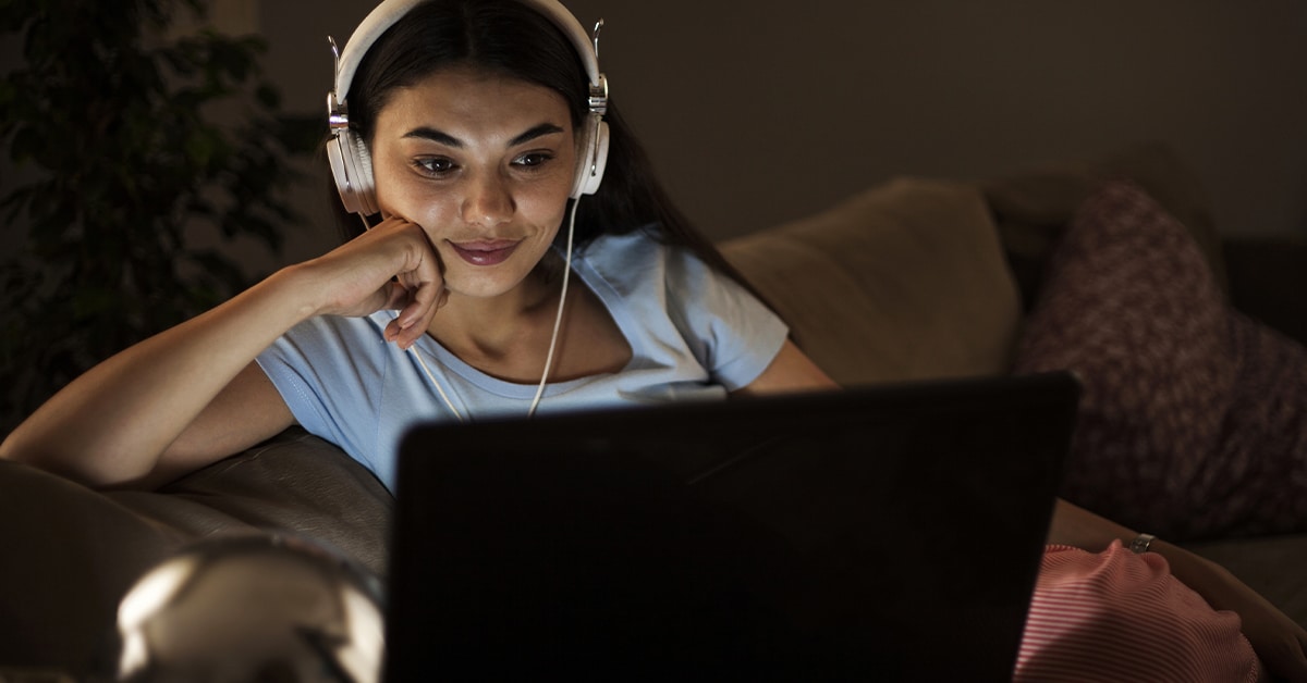 mulher negra de pele clara sentada em um sofá, com um notebook no colo. Ela, que veste blusa azul e calça listrada em branco e rosa, usa um headphone enquanto encosta o queixo em seu punho fechado. Seu antebraço, por sua vez, está apoiado no braço do sofá.