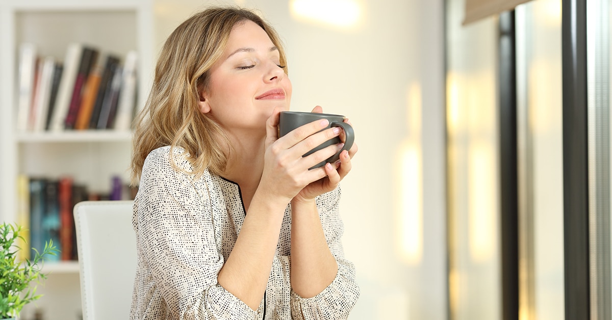 equilíbrio emocional: mulher branca segurando uma caneca, com expressão de quem sente um cheiro gostoso e rememora algo bom, prática de equilíbrio mental. Ao fundo, livros.