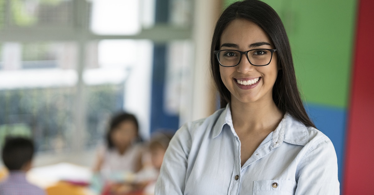 Pedadoga. Mulher parda sorri para a foto. Ela aparece da altura dos seios para cima, veste uma camisa jeans. Ao fundo, pessoas conversam em uma sala de aula.