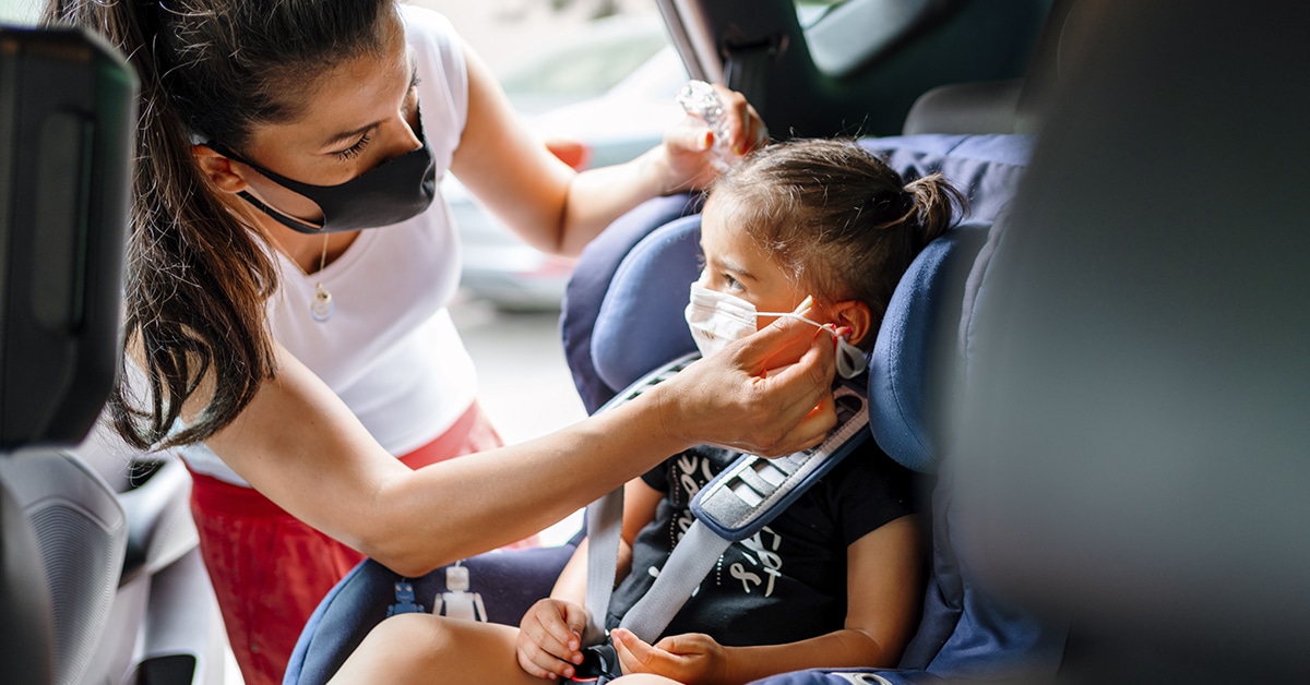 mulher arrumando criança em uma cadeirinha e seus dispositivos de retenção. A mulher está de fora do carro.