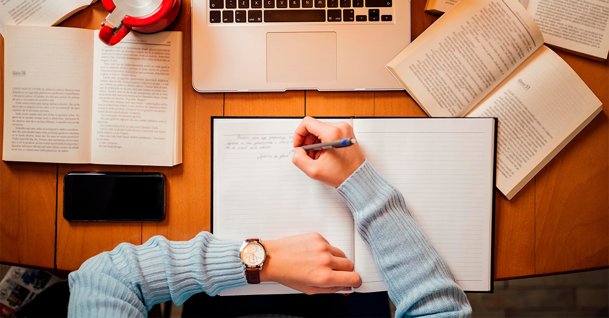 Pessoa com blusa azul segura caneta em uma das mãos em cima de um caderno onde escreve. Em cima da mesa há um celular a esquerda, livros abertos e um teclado de notebook.