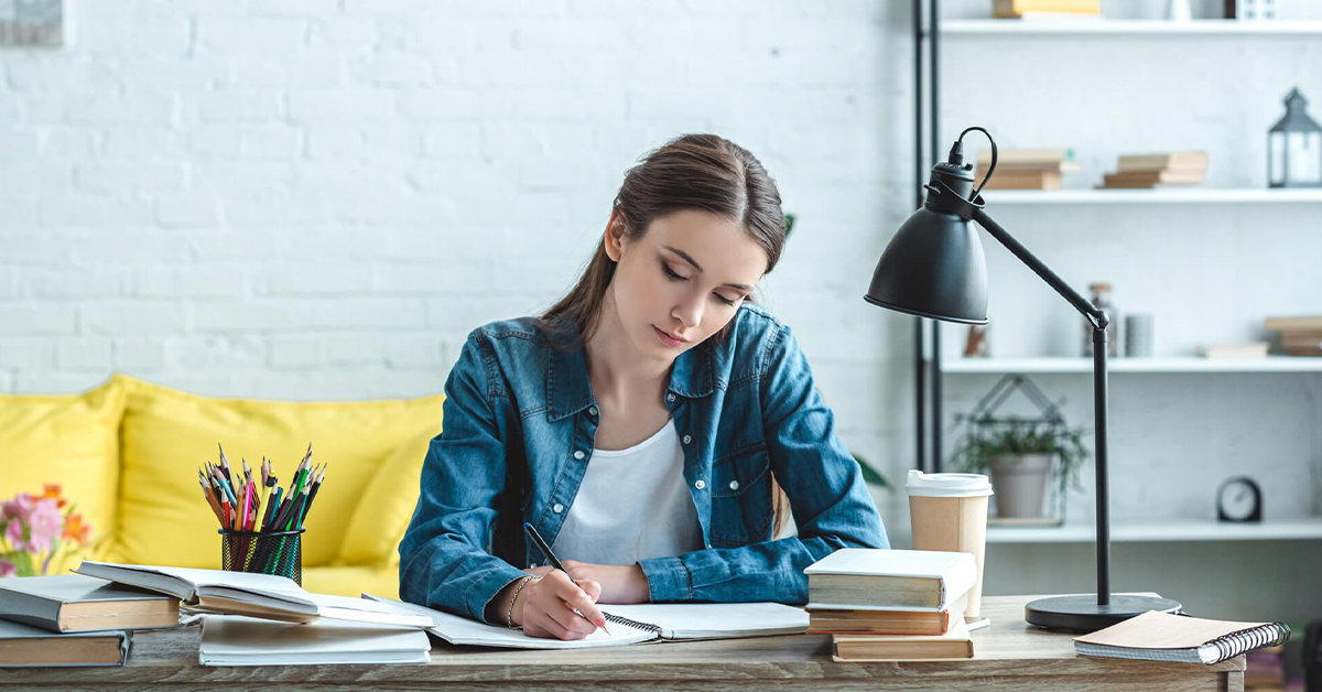 jovem mulher lendo um livro. ela está sentada, na mesa, a sua frente, há vários livros, várias obras e um abajur. Ao fundo, um sofá amarelo.