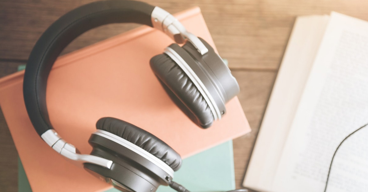 um headphone reto em cima de papeis rosa e azul em cima de uma mesa de madeira. Ao lado um livro aberto.