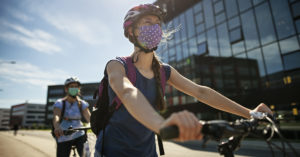 À frente mulher anda de bicicleta. Ela está com blusa cinz escuro, máscara e capacete. Atrás, homem anda de bicicleta. Ele está de camiseta, bermuda, máscara, óculos e capacete. Na lateral da extrema direita há uma grande prédio espelhado.