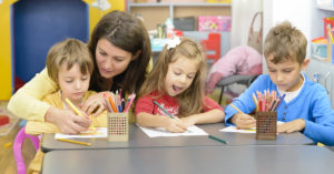a imagem mostra uma mulher e três crianças em uma sala de aula infantil. Ela está agachada e por trás da criança à esquerda, visto que a ajuda a segurar o lápis de cor amarelo.