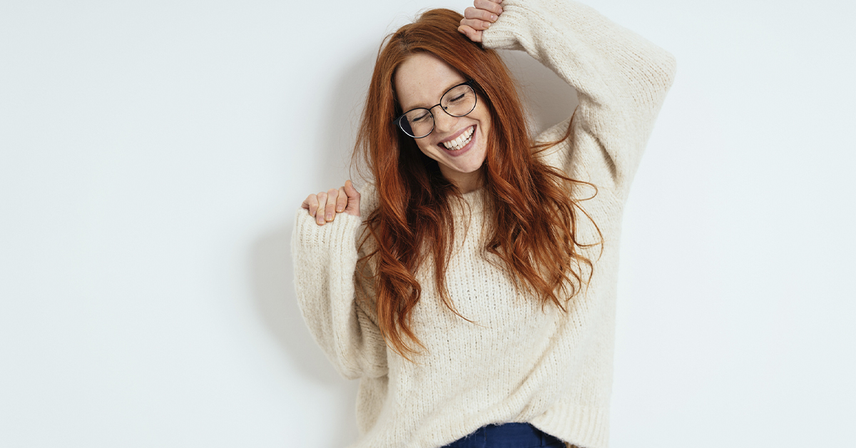 jovem branca ao centro da imagem. Ela é ruiva e posiciona seus braços para cima, seus punhos estão cerrados. Ela, que veste calça jeans e blusa de frio branca, sorri, mantendo os olhos fechados, como se comemora-se algo.