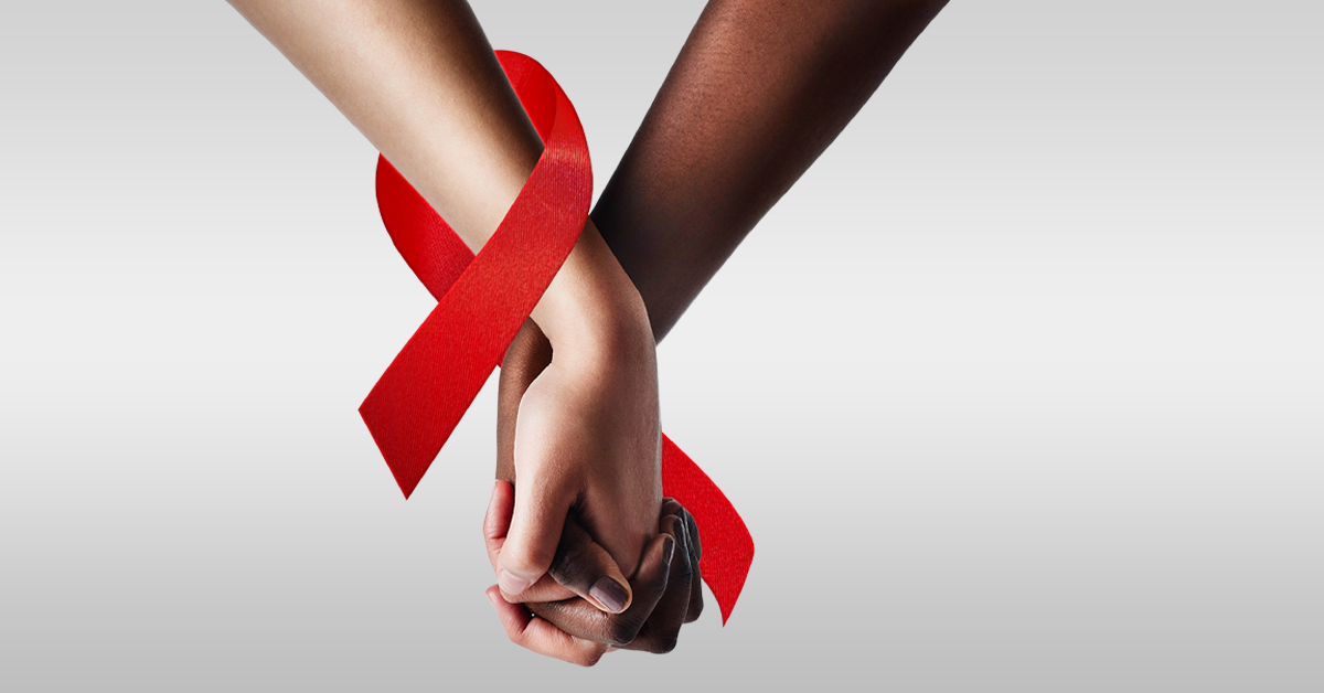 uma mão negra segura uma mão branca. Elas estão entrelaçadas com a fita vermelha que representa a luta contra a Aids.