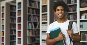 Estudante. Homem negro sorrindo veste blusa branca e carrega uma mochila nas costas. Ele está segurando materiais escolares, quanto segura, com a outra mão, a alça da mochila. Ao fundo, estantes repletas de livros.