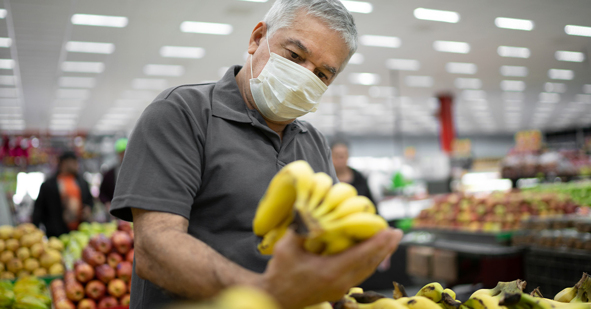 Homem de cabelo grisalho e pele clara, segurando bananas em um supermercado. Veste camiseta cinza escuro. Ao fundo desfocado supermercado e mulher em pé.